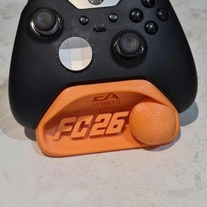 May include: Black Xbox controller resting on an orange stand. The stand has the text "EA SPORTS FC26" and a soccer ball design. The controller has black buttons and silver accents.
