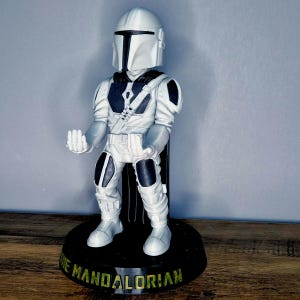May include: White and black Mandalorian figurine with a black base and the text "The Mandalorian" on the base.