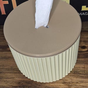 以下が含まれることがあります： ベージュのティッシュボックス、明るい茶色の蓋付き。円筒形の容器には、垂直方向のリブのディテールがあります。白いティッシュが蓋の中央から引き出されています。ティッシュボックスは木製の表面に置かれています。