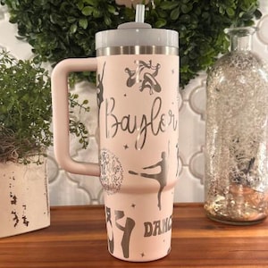 May include: A light pink tumbler with a gray lid and handle. The tumbler is decorated with silver ballet dancer silhouettes, stars, and the name "Baylor". The words "Dance" and "Just a girl who loves to dance" are also visible.