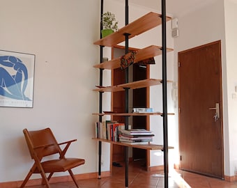 Könnte beinhalten: Ein brauner Ledersessel mit Holzarmlehnen und -beinen steht vor einem Holzregalsystem mit schwarzen Metallpfosten. Das Regalsystem hat fünf Regalbretter und ist mit Büchern, Pflanzen und anderen Gegenständen gefüllt. Das Regalsystem befindet sich in einem Raum mit einer weißen Decke und weißen Wänden. Eine braune Tür ist im Hintergrund sichtbar.