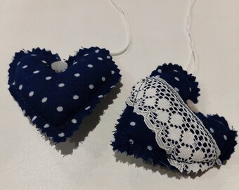 Puede incluir: Dos corazones de tela azul con lunares blancos y ribete de encaje. Un corazón tiene un botón blanco en el centro. Ambos corazones están unidos a cuerdas blancas.