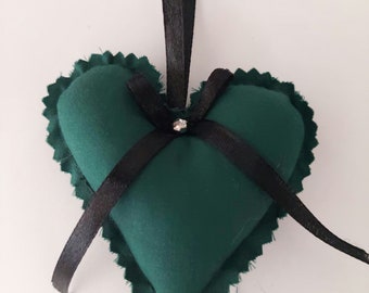 May include: A green fabric heart-shaped ornament with a black ribbon and a small rhinestone in the center. The ornament has a scalloped edge and is hanging from a black ribbon.