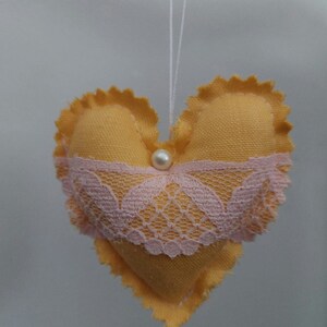 Könnte beinhalten: Handgefertigter herzförmiger Ornament aus gelbem Stoff mit gewelltem Rand. Ein rosa Spitzenband verläuft über die Mitte, und eine kleine Perle ist befestigt. Eine weiße Schnur ist zum Aufhängen angebracht.