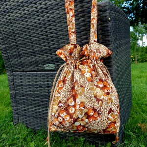 Puede incluir: Una bolsa de cordón de color marrón y naranja con estampado floral y una correa larga. La bolsa está colgada de una silla de mimbre.
