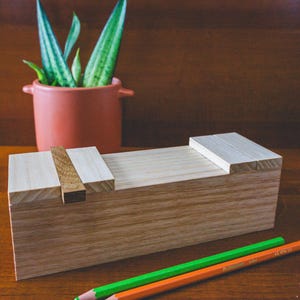 May include: A wooden pencil case with a sliding lid, in a natural wood finish. Two coloured pencils, one green and one orange, are placed on the surface. A terracotta pot with a green plant is in the background.