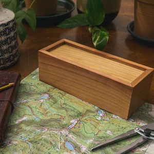 May include: A rectangular wooden box with a light brown finish and a sliding lid. The box is on a map with a compass and a leather-bound journal with a pencil. A decorative vase and potted plants are in the background.