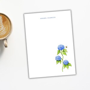 May include: White stationery with the name "ANNABEL HOLBROOK" in blue at the top. A watercolor illustration of blue hydrangeas and a bee is on the right side. A cup of coffee and a laptop are in the background.
