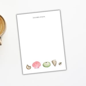May include: White stationery with the name "ANNABEL DAVIS" printed at the top. The bottom of the page features watercolor illustrations of seashells in shades of pink, green, and tan. A cup of coffee and a laptop are in the background.