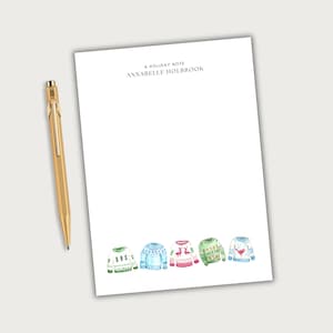 May include: A white rectangular notepad with the text A HOLIDAY NOTE and ANNABELLE HOLBROOK printed at the top, featuring a row of five colorful watercolor-style sweaters at the bottom, accompanied by a metallic gold-toned ballpoint pen.