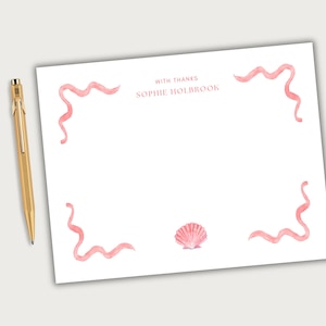 May include: White stationery with pink watercolor accents, including wavy lines and a seashell illustration. The text "WITH THANKS SOPHIE HOLBROOK" is printed in the upper center. A gold pen rests on the side.