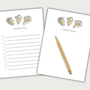 May include: Two white notepads with oyster illustrations and the text "CONRAD DAVIS". One notepad has a checklist, the other has a gold pen. The oyster illustrations are in shades of gray, yellow, and white.