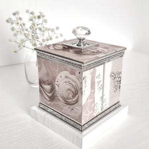 Peut inclure: Une boîte carrée décorative avec un motif floral de roses dans des tons de rose et de crème. La boîte est ornée d'un bouton en cristal et de bordures argentées et de strass. La boîte repose sur une base blanche.