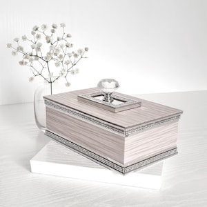 Peut inclure: Boîte rectangulaire décorative avec un effet de grain de bois clair, garniture argentée et détails en cristal. Elle est ornée d'un bouton en cristal et repose sur une base blanche. Un vase de fleurs blanches est en arrière-plan.