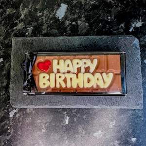 May include: A chocolate bar with the words "Happy Birthday" in white chocolate letters and a red heart on a slate background.