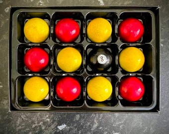 Può includere: Un vassoio nero con 12 caramelle rotonde di cioccolato. Le caramelle sono disposte in una griglia 3x4. Le caramelle sono gialle, rosse e nere. La caramella nera ha il numero 8.