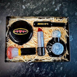 May include: A set of black and silver makeup items in a box with shredded paper. The set includes a compact with hearts, lipstick, mascara, perfume bottle, and a makeup brush.