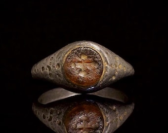 May include: A weathered silver-toned metal ring featuring a circular, amber-colored stone inlay at the center. The surface of the metal band displays a textured, pitted appearance, and the stone is engraved with a subtle cross-like symbol.