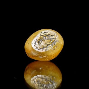 Puede incluir: Un sello de piedra de cornalina amarilla con un diseño tallado de un animal estilizado, posiblemente un pájaro o una criatura mítica. El sello está tallado en un estilo detallado e intrincado.