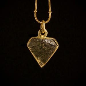 Può includere: Una collana con pendente dorato a forma di diamante, con una pietra scura. Il pendente è sospeso a una sottile catena d'oro su sfondo nero. La collana è un gioiello.