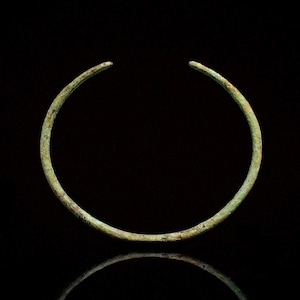 May include: A circular open-ended bracelet made of weathered bronze metal with a textured, greenish-brown patina. The band features a simple, uniform thickness throughout its circumference, displaying signs of age and oxidation on its surface.