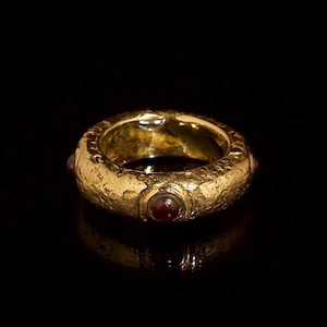 Puede incluir: Un anillo de banda gruesa en oro con una superficie texturizada y martillada. El anillo presenta varias piedras preciosas pequeñas, redondas y de color rojo oscuro incrustadas a intervalos regulares alrededor de la banda metálica.