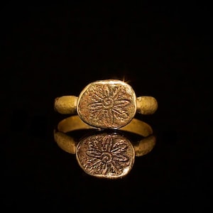May include: A gold ring featuring a floral pattern on its face. The ring has a rectangular, rounded face and a smooth band. The ring's reflection is visible on the dark surface beneath it.