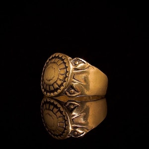 May include: A gold-toned metal ring featuring a circular face with a sunburst-like radial pattern of etched lines and a scalloped border. The band displays decorative engraved details on the sides, including small triangular and dot motifs.