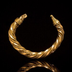 May include: A gold-colored, twisted metal bracelet with detailed dragon head terminals. The bracelet's design features a braided pattern and is set against a black background, with its reflection visible below.