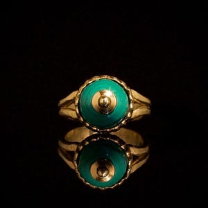 Può includere: Un anello d'oro con una pietra rotonda color verde acqua e un centro dorato. L'anello ha una fascia in oro strutturata e un'incastonatura smerlata attorno alla pietra. L'anello è esposto su una superficie nera riflettente.