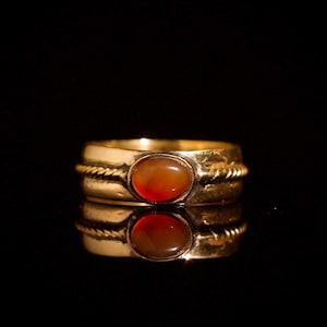 Könnte beinhalten: Ein goldener Ring mit einem ovalen, rot-orangen Stein. Der Ring hat ein dickes Band mit einem seilartigen Design um den Stein. Der Ring spiegelt sich auf einer dunklen, glänzenden Oberfläche.