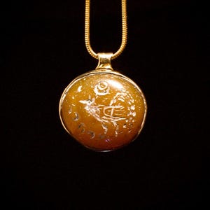Peut inclure: Un collier en chaîne dorée avec un pendentif rond de couleur ambre orné d'une gravure représentant un oiseau avec une lune croissante et d'autres symboles.