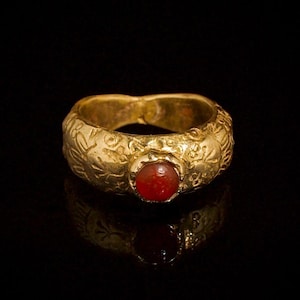 May include: A gold ring with a red gemstone set in the center. The ring has intricate embossed designs around the band. The ring is displayed against a black background, with its reflection visible.