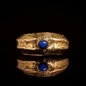 May include: A wide gold-toned ring with a textured, hammered surface and a central oval-shaped blue stone. The band features a rugged, uneven finish that contrasts with the smooth, polished appearance of the small, deep blue cabochon gemstone.
