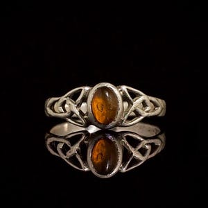 Könnte beinhalten: Ein silberfarbener Ring mit einem ovalen, durchscheinenden bernsteinfarbenen Stein in der Mitte. Die Ringschiene ist mit einem komplizierten, durchbrochenen Knotenmuster gestaltet, das sich von der zentralen Fassung zu beiden Seiten erstreckt.