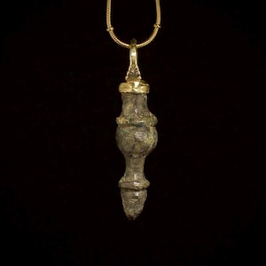 Puede incluir: Un collar con colgante que presenta un colgante cilíndrico ornamentado de color bronce, con una tapa y un asa de color dorado. El colgante cuelga de una delicada cadena dorada sobre un fondo negro.