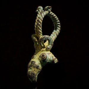 Pode incluir: Pingente antigo em bronze, com cabeça de veado e design de corda em laço. O pingente tem uma aparência envelhecida com pátina verde. O olho do veado é um pequeno detalhe circular. O fundo é preto, destacando os detalhes.