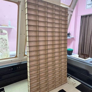 May include: A tall, rectangular wooden display rack featuring a grid of numerous small, open compartments arranged in vertical columns and horizontal rows. The structure is constructed from light-colored plywood with visible dark edges.