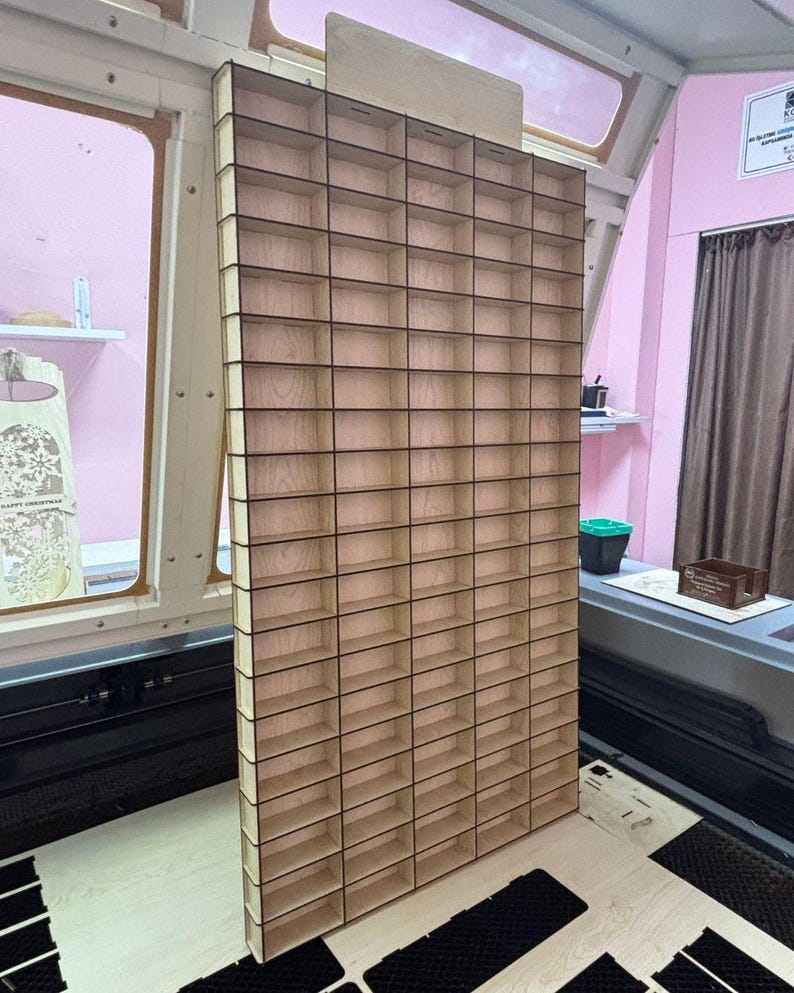 May include: A tall, rectangular wooden display rack featuring a grid of numerous small, open compartments arranged in vertical columns and horizontal rows. The structure is constructed from light-colored plywood with visible dark edges.
