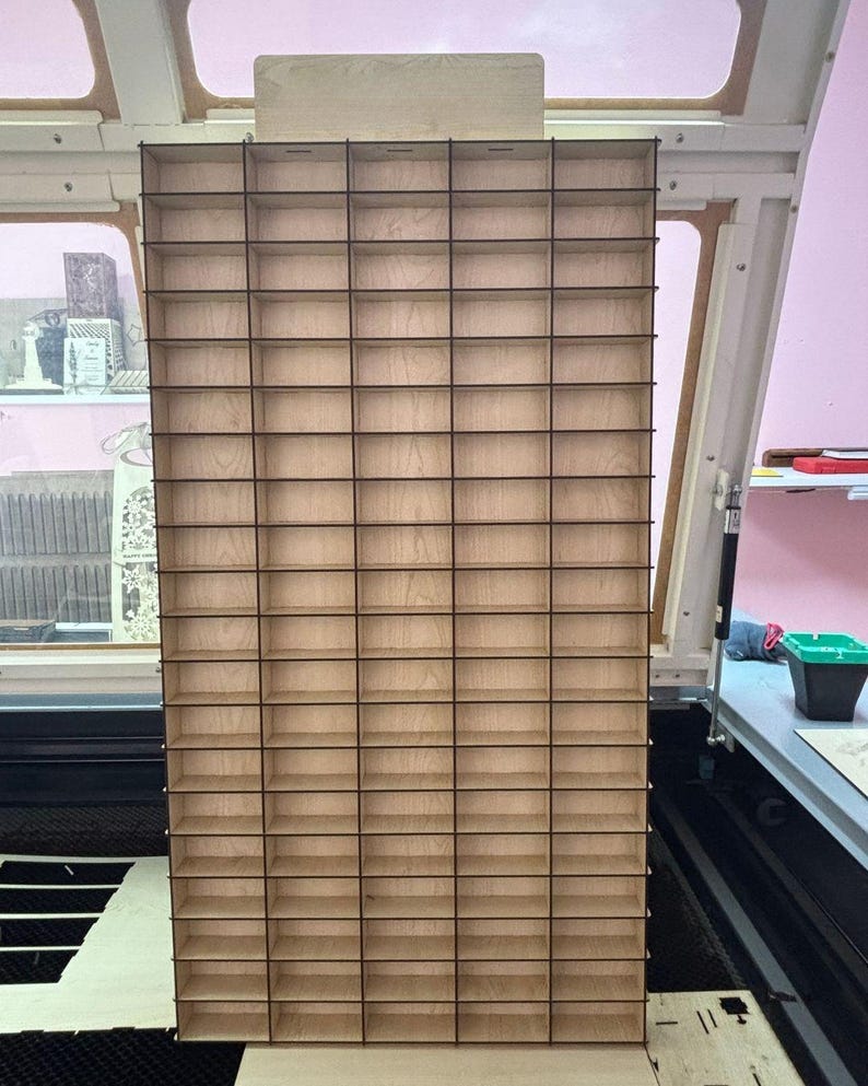 May include: A tall, rectangular wooden storage organizer featuring a grid of numerous small, open compartments. The structure is constructed from light-colored plywood with dark, thin dividers creating a uniform pattern of rectangular cubbies.