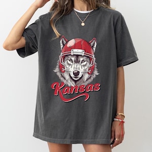 May include: Gray oversized t-shirt with a graphic of a wolf wearing a red football helmet. The text "Kansas" is printed in red below the wolf.