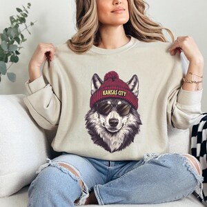May include: Beige sweatshirt featuring a graphic of a wolf wearing a maroon beanie with "KANSAS CITY" printed on it and sunglasses. The wolf is depicted in a detailed, realistic style. The sweatshirt has long sleeves and a crew neck.