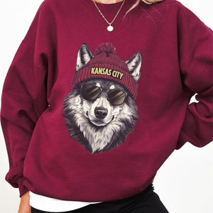 May include: Burgundy sweatshirt featuring a smiling wolf wearing sunglasses and a red beanie with "KANSAS CITY" in yellow lettering. The wolf has black and white fur. The sweatshirt has long sleeves.