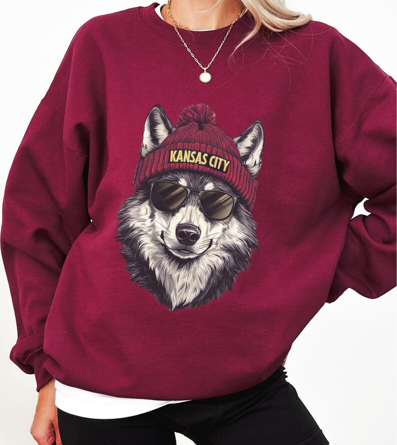 May include: Burgundy sweatshirt featuring a smiling wolf wearing sunglasses and a red beanie with "KANSAS CITY" in yellow lettering. The wolf has black and white fur. The sweatshirt has long sleeves.