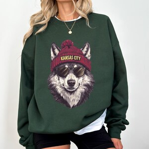 May include: A dark green sweatshirt featuring a wolf wearing a red beanie with "KANSAS CITY" printed on it and sunglasses. The wolf graphic is centered on the front of the sweatshirt. The sweatshirt has long sleeves and a crew neck.