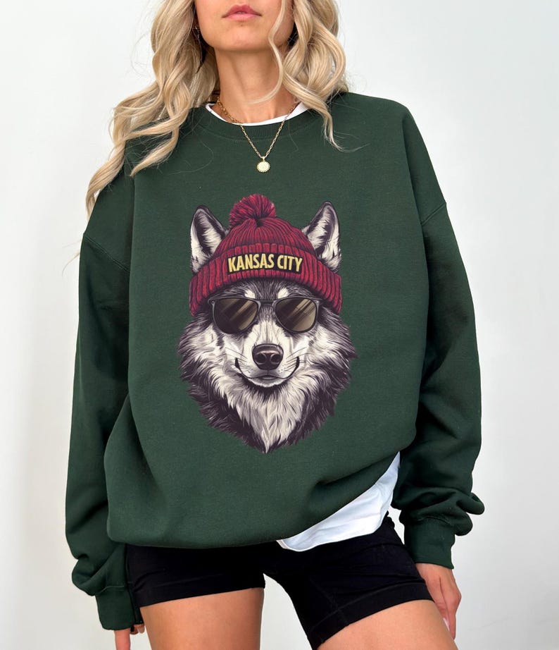 May include: A dark green sweatshirt featuring a wolf wearing a red beanie with "KANSAS CITY" printed on it and sunglasses. The wolf graphic is centered on the front of the sweatshirt. The sweatshirt has long sleeves and a crew neck.