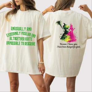 May include: A white t-shirt with green text that reads "Unusually and Exceedingly Peculiar and Altogether Quite Impossible to Describe". The back of the shirt features a watercolor illustration of two women, one in green and one in pink, with the text "Because I knew you, I have been changed for good."