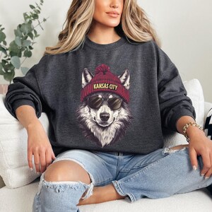 May include: Dark gray sweatshirt featuring a wolf wearing sunglasses and a maroon beanie with "Kansas City" text. The wolf graphic is detailed with shades of gray and white. A casual, comfortable top.