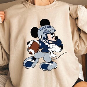 May include: A beige sweatshirt with a cartoon image of Mickey Mouse wearing a football helmet and jersey. The jersey has the text "Dallas Est 1960" on it. Mickey is holding a football and is in a running pose.