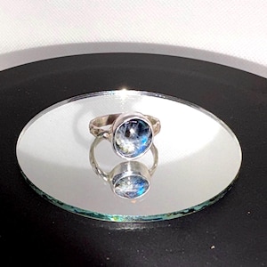 May include: A silver ring featuring a round, cabochon-style stone with a dark, iridescent surface displaying flashes of blue and white light. The metal band has a textured, slightly irregular finish, and the stone is set in a smooth silver bezel.